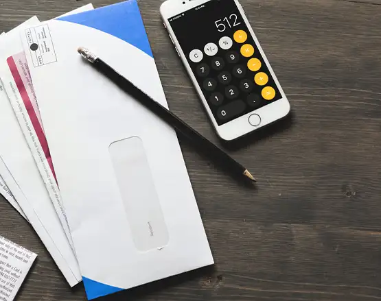 Smartphone with calculator app open next to pen, envelopes, and paperwork on wooden desk