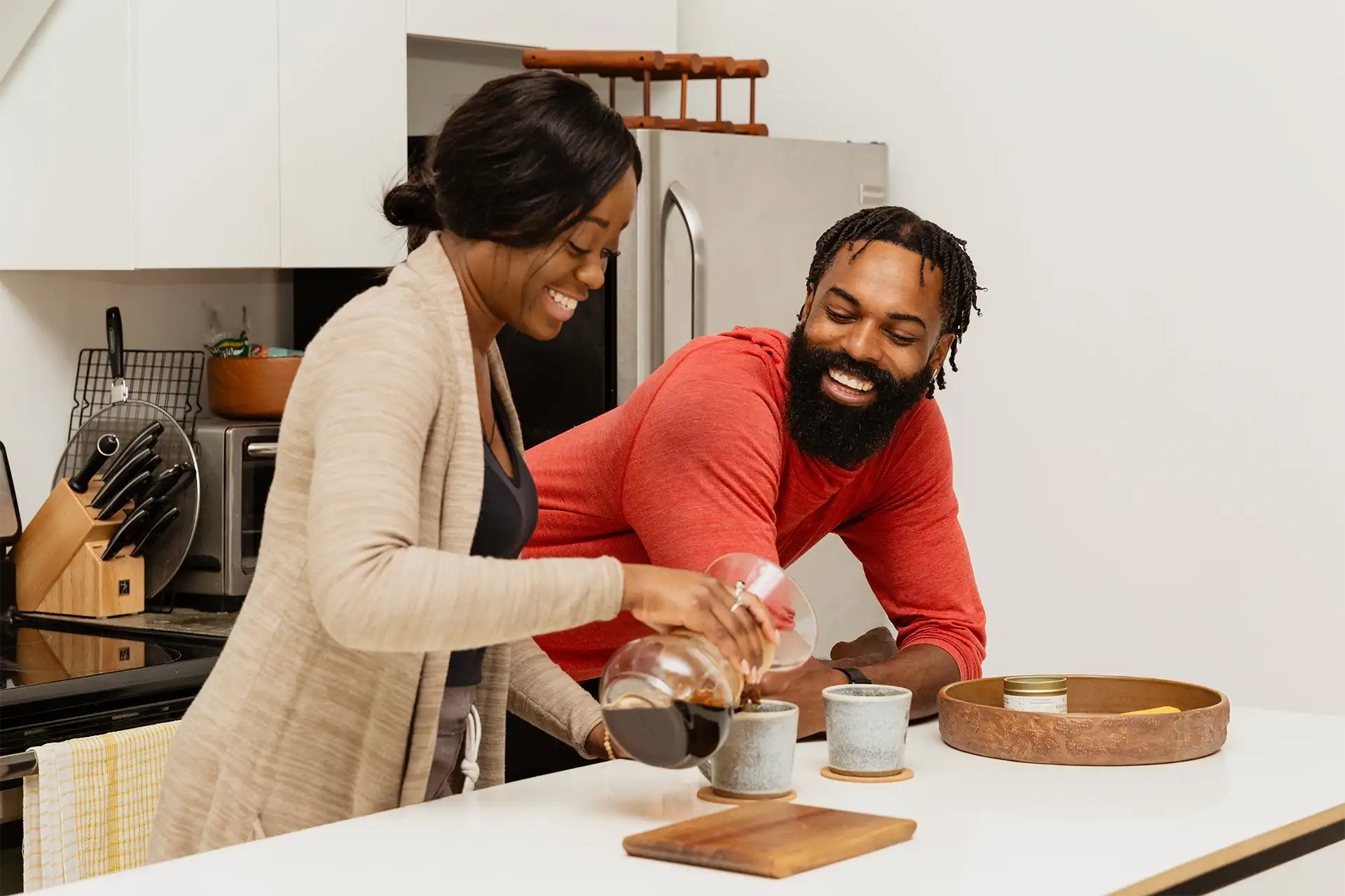 Smiling couple preparing pour-over coffee together in modern kitchen with ceramic mugs and wooden tray
