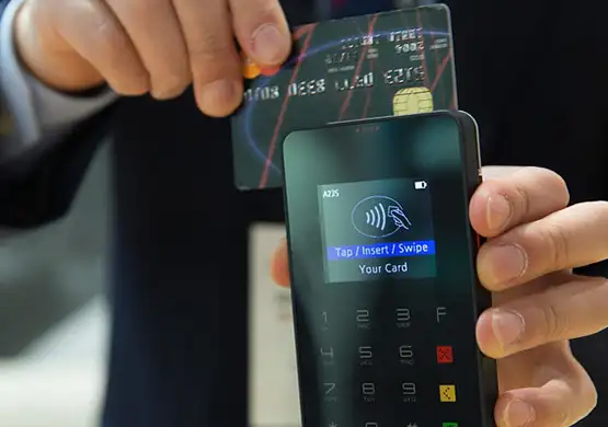 Hand holding credit card above payment terminal displaying contactless tap to pay symbol and keypad