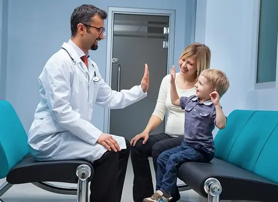 Doctor in white coat giving high-five to young boy on couch while mother watches in medical office