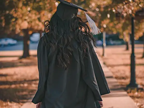 Graduate in black cap and gown walking outdoors in autumn with fallen leaves on ground