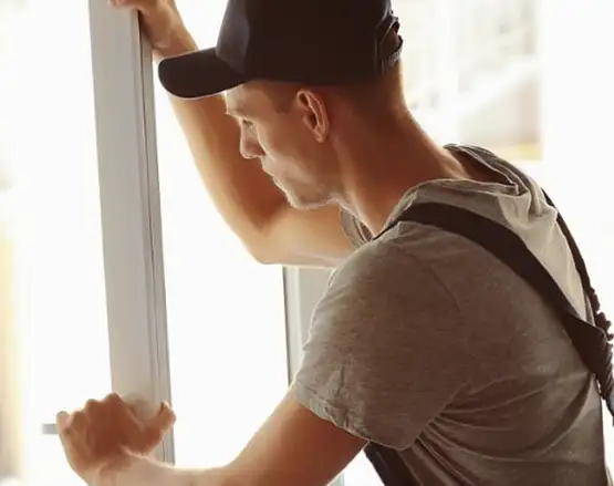 Man in baseball cap and backpack installing or measuring a white window frame indoors