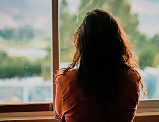 Woman with long dark hair looking out window at blurred green landscape while wearing rust-colored sweater