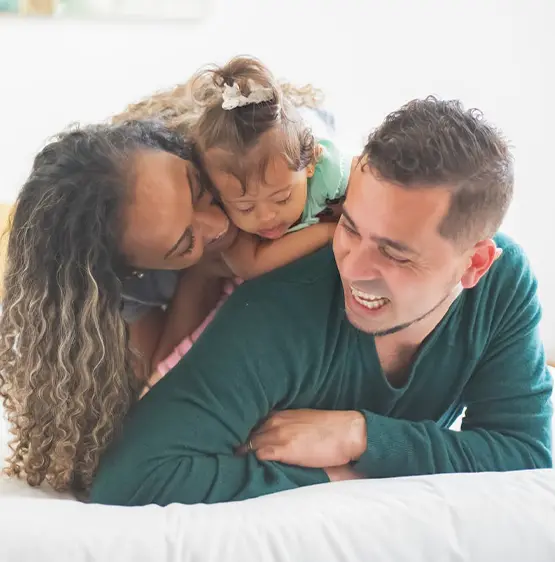 Happy family of three lying on bed together, with parents smiling at their young daughter between them