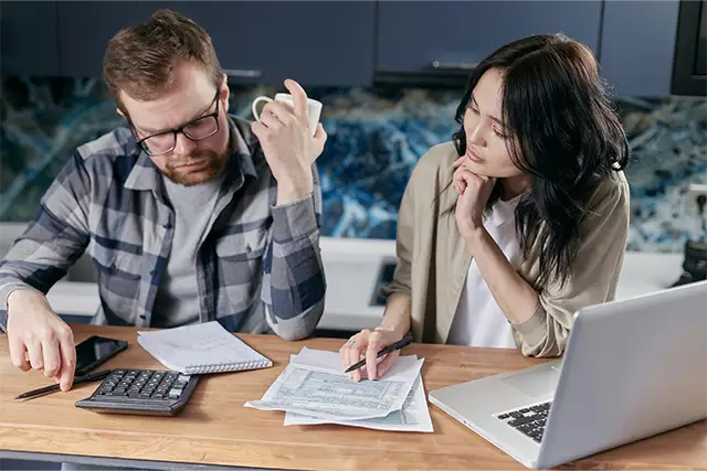 Couple reviewing financial documents with calculator and laptop at home table looking concerned