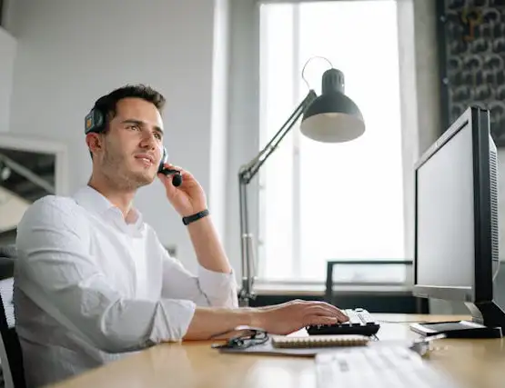 Professional man in white shirt talking on phone while working at computer desk in bright office