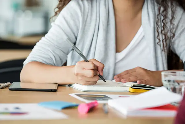 Woman writing notes at desk with pen and paper surrounded by colorful office supplies and sticky notes