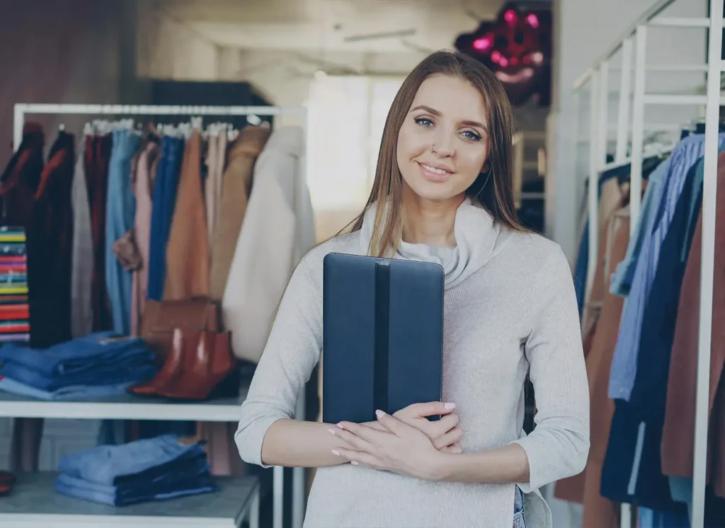 Woman in beige sweater holding tablet in clothing boutique with racks of apparel
