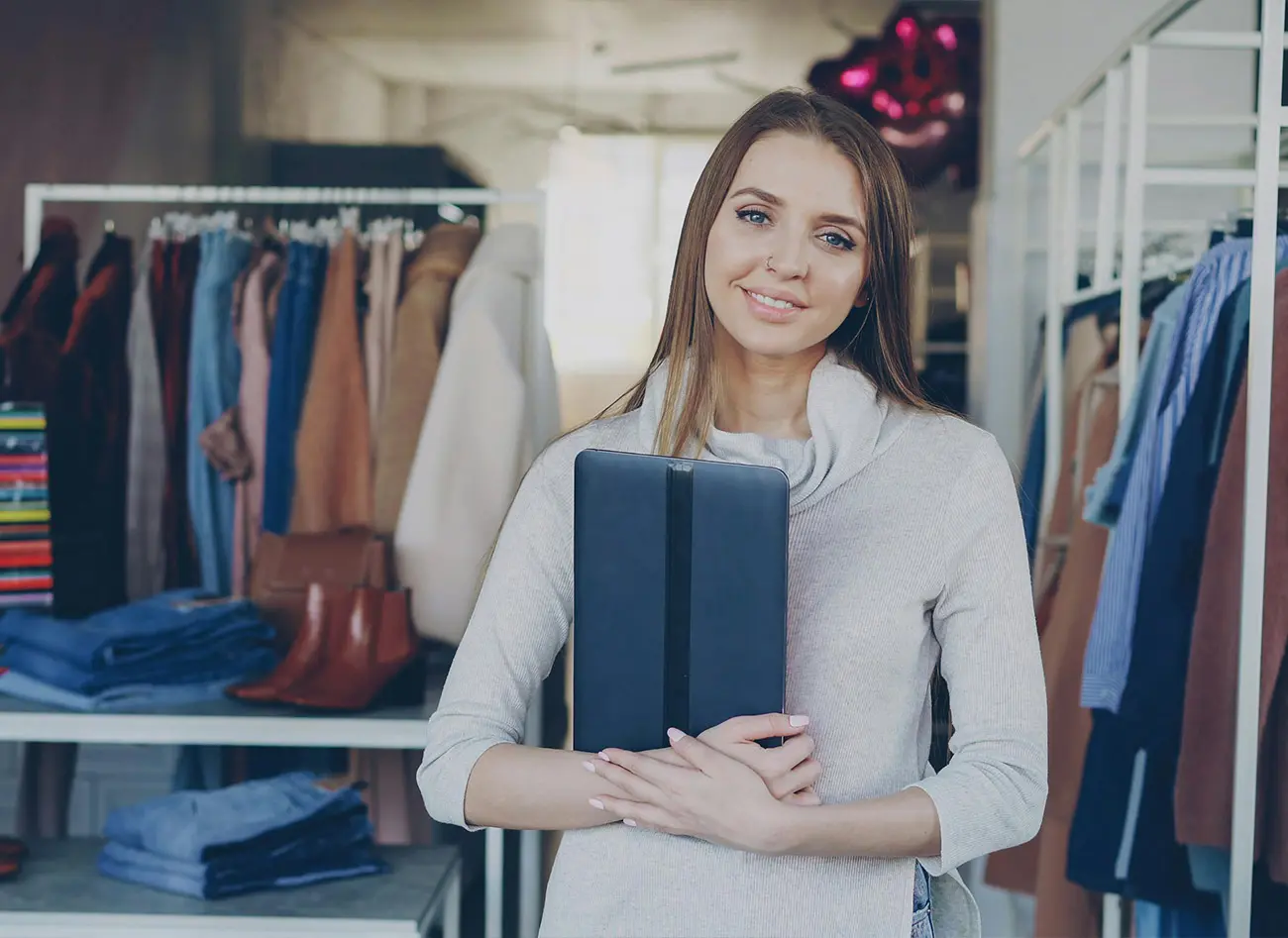 Woman in beige sweater holding tablet in clothing boutique with racks of apparel