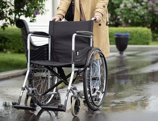 Person in tan coat pushing empty wheelchair on wet pavement outdoors