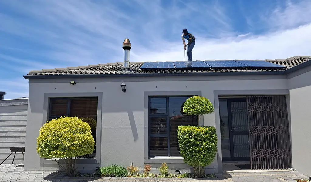 Technician installing solar panels on residential roof with trimmed bushes and gray exterior walls below