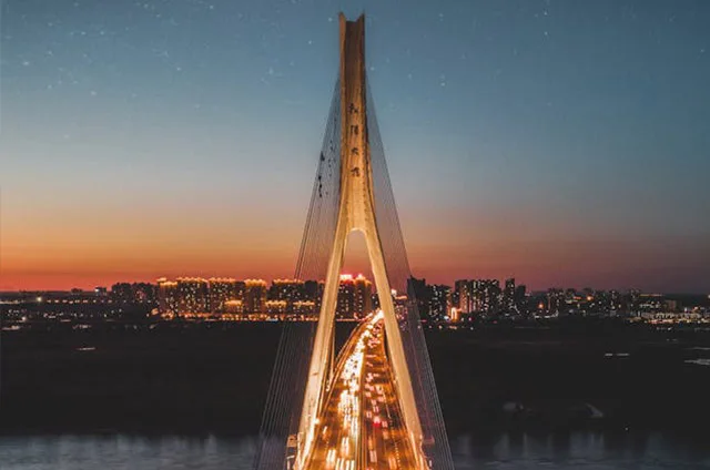 Illuminated cable-stayed bridge spanning water at sunset with city skyline in background