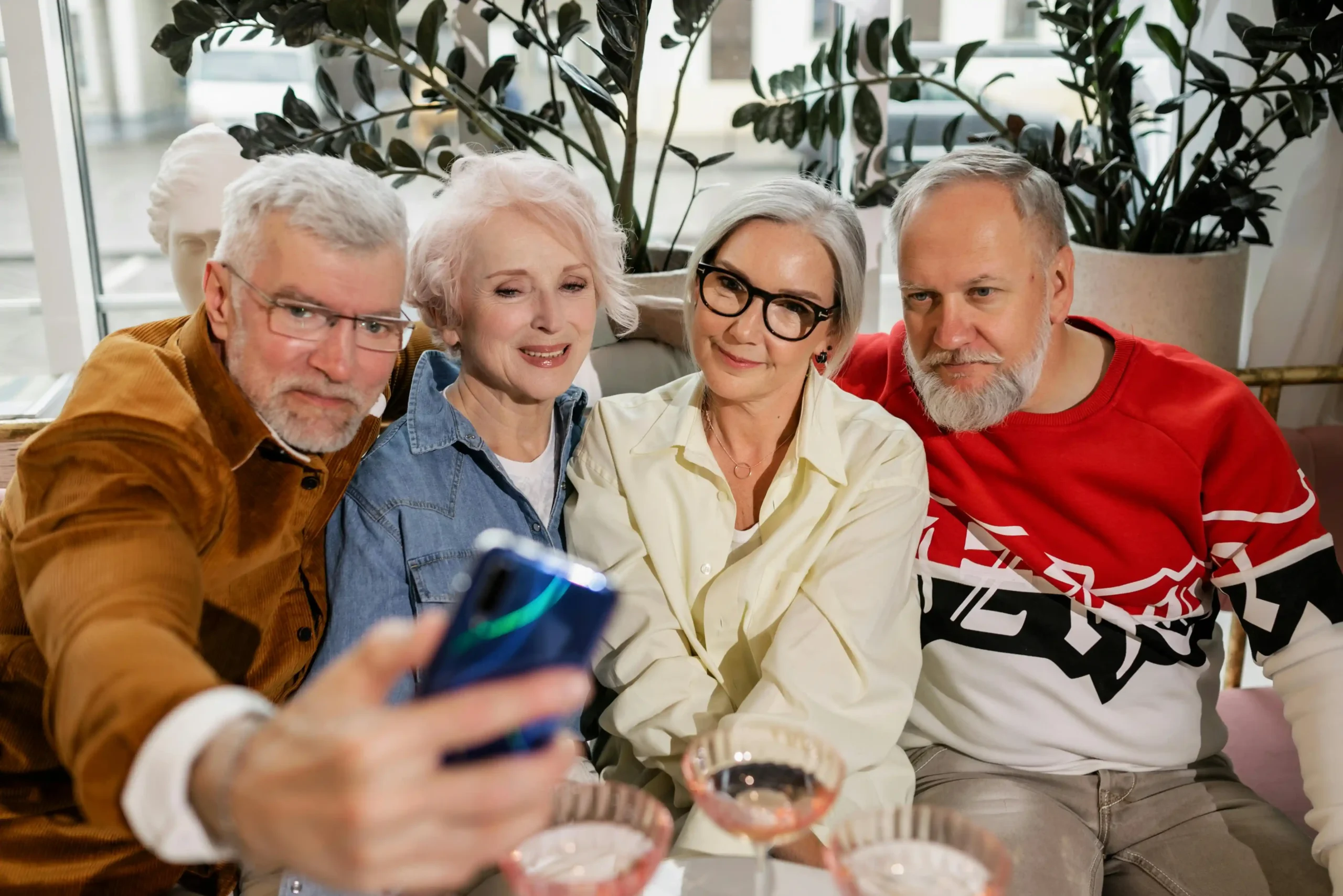 Four senior friends taking a selfie together on a couch in a bright, plant-filled living room