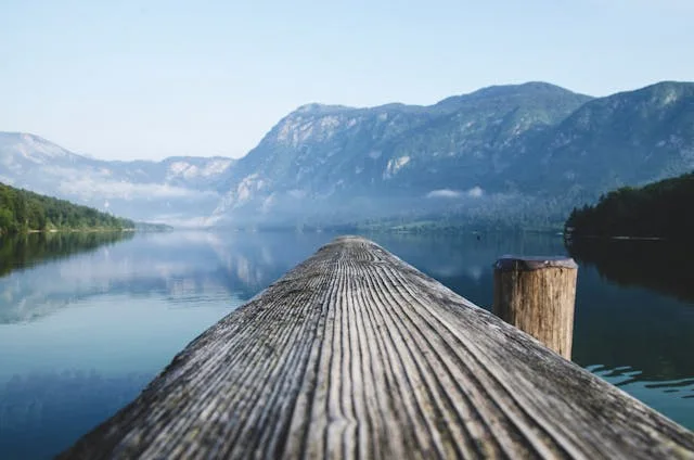 Wooden dock extending over calm mountain lake with forested shores and misty peaks reflected in water