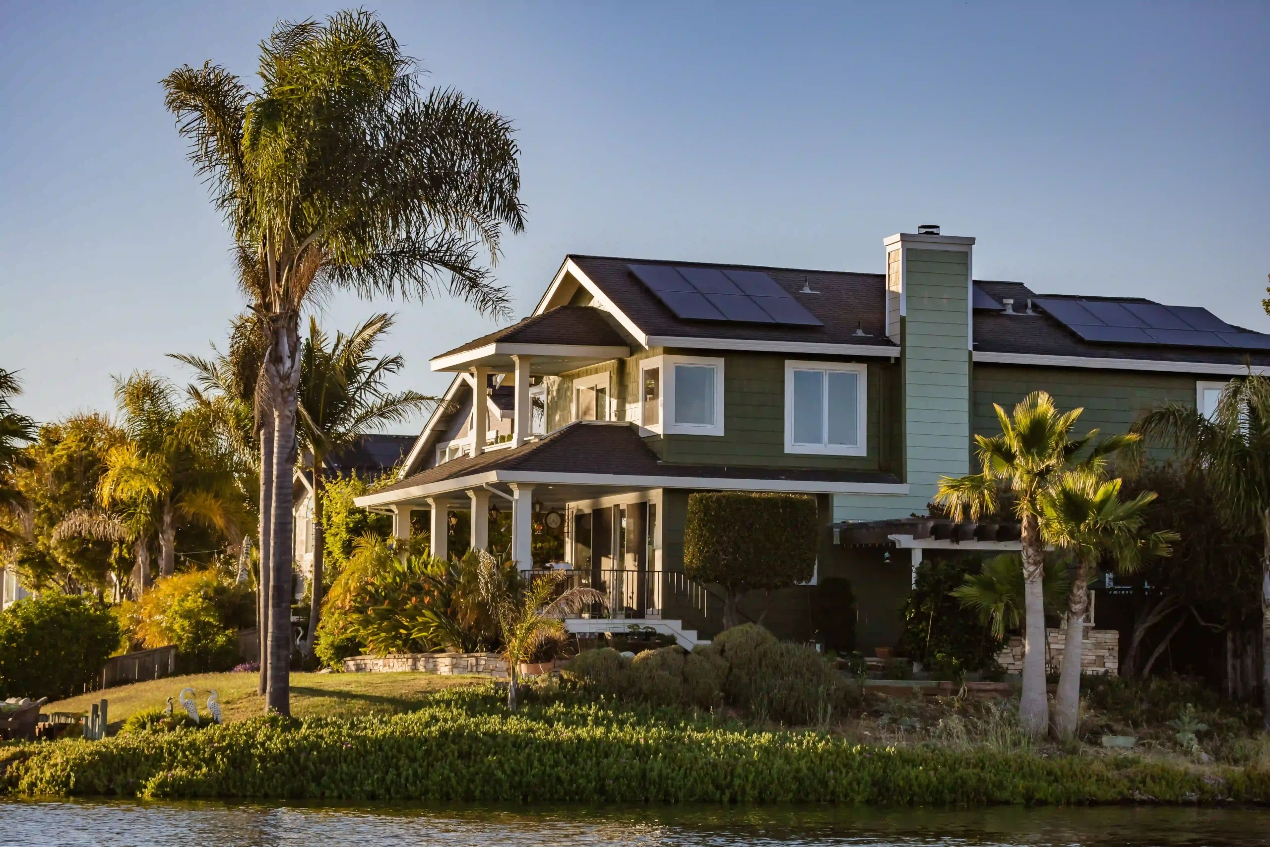 Two-story craftsman home with solar panels on roof, palm trees, and waterfront landscaping at sunset